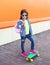 Fashion little girl child with skateboard wearing a sunglasses and checkered shirt and backpack over orange