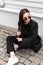 Fashion lovely young woman in stylish sunglasses in fashionable black clothes from new summer trendy collection is sit on stone