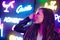 Fashion portrait of a young woman in sunglasses posing near neon signs in night club