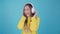 Fashion pretty cool smiling girl listening to music in headphones wearing yellow sweater over blue background . 4k