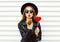 Fashion pretty sweet young woman with red lips sends air kiss with lollipop heart wearing black hat leather jacket over white