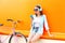 Fashion pretty young woman listens to music using smartphone near urban retro bicycle over colorful orange