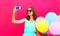 Fashion smiling woman taking a picture on a smartphone with an air colorful balloons on pink background