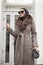 Fashion woman in the city stands in front of a white door. She is wearing a coat. sunglasses and bag