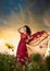 Fashionable beautiful young woman in long red dress posing outdoor with cloudy dramatic sky in background. Attractive brunette