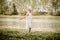 Fashionable child in white dress dancing in grass by the river and posing in summer time