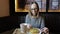 Fashionable girl in glasses with a smile eating pankcakes with syrup and drinking herbal tea sitting in a restaurant.