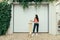 Fashionable girl in street clothes stands background of white garage door with longboard in her hands and looks away.Stylish