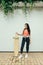 Fashionable girl in street clothes stands background of white garage door with longboard in her hands and looks down.Stylish