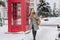 Fashionable joyful young woman with long brunette hair walking on street full with snow on red telephone box background