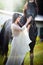 Fashionable lady with white bridal dress near brown horse. Beautiful young woman in a long dress posing with a friendly horse