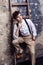 Fashionable man in white shirt and beige pants with suspenders sitting and posing on wooden ladder on brick wall background