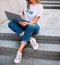 Fashionable woman sitting on stairs with laptop, wearing ripped jeans,white T-shirtand sneakers. fashion blogger outfit details