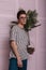 Fashionable young man in trendy clothes with glasses poses with an exotic palm tree in a pot near wall. Handsome hipster guy in