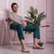 Fashionable young man in trendy clothes with glasses sits on a chair near an exotic palm tree in a pot. Handsome hipster guy in