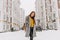 Fashionable young woman in coat with backpack walking on street in big city in snowing time. Cheerful mood, snowfall