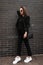 Fashionable young woman in stylish jeans trousers in long vintage blazer with leather black trendy handbag posing near brick wall