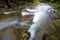 Fast flowing through wild green forest river with crystal clear smooth silky water falling from big wet stones in beautiful