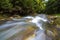 Fast flowing through wild green forest river with crystal clear smooth silky water falling from big wet stones in beautiful