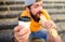 Fast food meal for lunch. Hipster bite hot dog hold drink paper cup. Man bearded shows paper cup drink stairs background