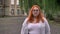 Fat ginger girl with glasses is standing outside in park in gloomy weather, watching at camera