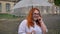 Fat ginger girl with glasses is standing under rain in park, talking on phone, holding umbrella, communication concept