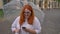 Fat ginger girl with glasses is typing message on smartphone in park in rainy weather, laughing, holding umbrella