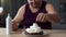 Fat male putting cherry on the top of sweet cake decorated with whipped cream