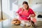 Fat young woman in kitchen sitting and eating sweet food. Confused serious plus size model look on sweets. Body positive