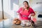 Fat young woman in kitchen sitting and eating sweet food. Soft tape measure around waist and in hands. Plus size model