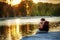 Father and 2 years old child play fishing on bank of lake in sunset time. Golden in sun light trees and water around. Father and