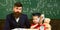 Father with beard, teacher teaches son, kid boy. Teacher and pupil in mortarboard, chalkboard on background. Kid studies