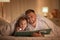 Father and daughter with flashlight reading book under blanket