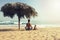 Father and his toddler son practicing yoga at the beach. They sits in pose of lotus under big cane sun umbrella and looks at the