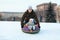 Father push her child from snowy slope, kid sitting in an inflatable toboggan, winter season is in city