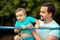 Father supporting toddler son doing muscle up exercise on steel bar. Dad and little son training together outdoors in summer