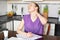 Fatigue woman feels tired of sitting long time at kitchen table, keeps hand on neck, dressed in purple t shirt, writes in papers,