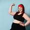 Fatty ginger lady in red headband, black top and leggings, fitness bracelet. Showing her muscles, smiling. Blue background