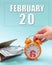 February 20th. Hand holding an orange alarm clock, a wallet with cash and a calendar date. Day 20 of month.