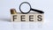 The FEES sign on wooden block on white background with coins and magnifier