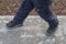 Feet of boy dressed in dark pants, black boots, walking on snow-covered parapet rising on blurred background of fallen foliage