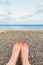 Feet with a red pedicure on the background of a beach made of pebbles and small seashells with the sea.