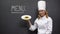 Female chief cook holding pasta plate with menu word on blackboard, restaurant