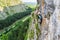 Female climber switching carabiners on a via ferrata route, vertical wall high above the valley, with a road behind.