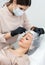 Female cosmetologist or dermatologist making a patient facial procedure in the cosmetology office. Beautician applying face mask