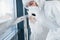 Female doctor scientist in lab coat, defensive eyewear and mask standing indoors with antibacterial spray