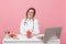 Female doctor sit at desk work on computer with medical document hold angel in hospital isolated on pastel pink wall