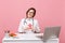 Female doctor sit at desk work on computer with medical document hold angel in hospital isolated on pastel pink wall