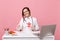 Female doctor sit at desk work on computer with medical document hold angel in hospital isolated on pastel pink wall