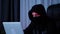 Female face in black mask and hoodie looking at laptop screen. Hacker face in black hood looking at computer and typing code. Woma